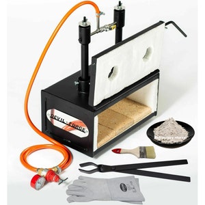 May include: A black metal gas forge with a white insulated door, a red pressure gauge, and a set of black tongs. The forge is labelled "DEVIL-FORGE" and includes a bag of refractory mortar.