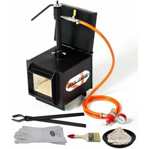 May include: A black metal forge with an orange hose and a red and white label that says "Devil-Forge". The forge is on a white background and includes a pair of tongs, a pair of leather gloves, a paintbrush, and a small plate of refractory mortar.