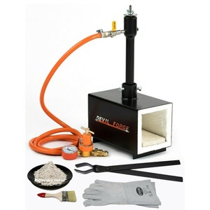 Puede incluir: Un horno de metal negro con una manguera naranja y un regulador. El horno tiene un revestimiento refractario blanco y el texto "DEVIL FORGE" está impreso en el lateral. El conjunto incluye unas tenazas, un pincel, una bolsa de mortero refractario y un par de guantes de soldadura.