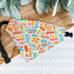 May include: A white bandana with a colorful print of empowering words like "love", "power", "brave", and "believe". The bandana has a black adjustable strap.