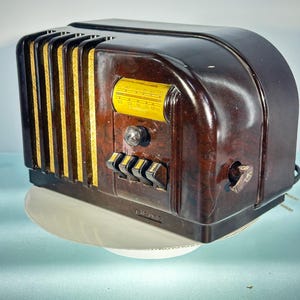 May include: A vintage brown radio with a curved top and vertical gold-coloured accents. The radio features a yellow dial with frequency markings and several black control buttons. A power cord extends from the side.