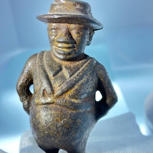 Peut inclure: Une figurine en fonte d'une personne portant un chapeau et une veste. La figurine est peinte en brun foncé et a une surface texturée.