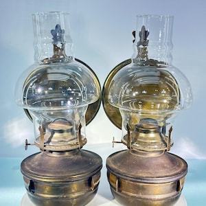 May include: Two antique oil lamps with clear glass globes and chimneys. The lamps have brass bases and accents, and are mounted on circular brass backplates. The lamps are designed to be mounted on a wall.