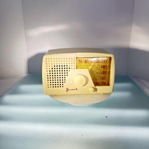Peut inclure: Une radio AM vintage de marque Jewel de couleur crème avec une grille de haut-parleur noire et un cadran rouge et jaune. La radio a un bouton de réglage et un logo Jewel rouge.