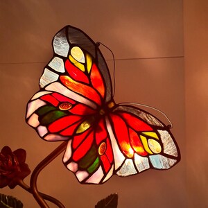 May include: A stained glass butterfly lamp with a red, yellow, and blue design. The butterfly is perched on a metal stem with a rose.