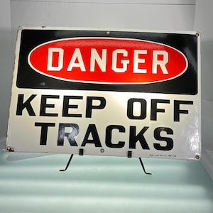 May include: A white and black enamel sign with a red oval that says "DANGER" in white letters. The sign also says "KEEP OFF TRACKS" in black letters. The sign is made by Ready Made Sign Co. New York.