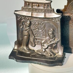 May include: A pair of antique bronze bookends depicting a Japanese scene with a woman in a kimono and a man playing a musical instrument. The bookends are shaped like a pagoda with intricate details.