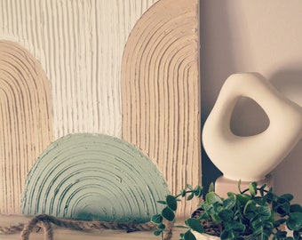 May include: A textured wall art piece with a modern abstract design featuring beige, white, and green arches. The art is displayed on a shelf with a white ceramic sculpture and a potted plant.