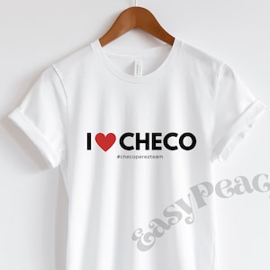 Könnte beinhalten: Weißes T-Shirt mit dem Text "I ❤️ CHECO" in Schwarz und Rot. Unter dem Text befindet sich der Hashtag "#checoperezteam". Das T-Shirt hängt an einem Holzbügel.