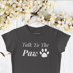 Op de afbeelding: Een donkergrijs T-shirt met korte mouwen met de tekst "Talk To The Paw" en een witte voetafdruk.