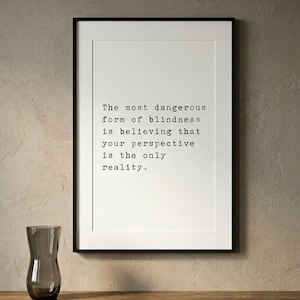 May include: A framed wall art print featuring the black text: The most dangerous form of blindness is believing that your perspective is the only reality. The print is displayed in a thin black frame with a white mat border on a textured wall.