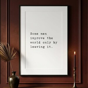 May include: A rectangular white wall art print in a thin black frame featuring the black text: Some men improve the world only by leaving it. The print is displayed on a dark red panelled wall above a wooden console table with a vase and candle.