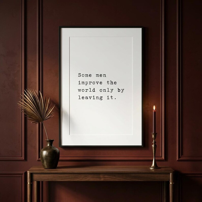 May include: A rectangular white wall art print in a thin black frame featuring the black text: Some men improve the world only by leaving it. The print is displayed on a dark red panelled wall above a wooden console table with a vase and candle.