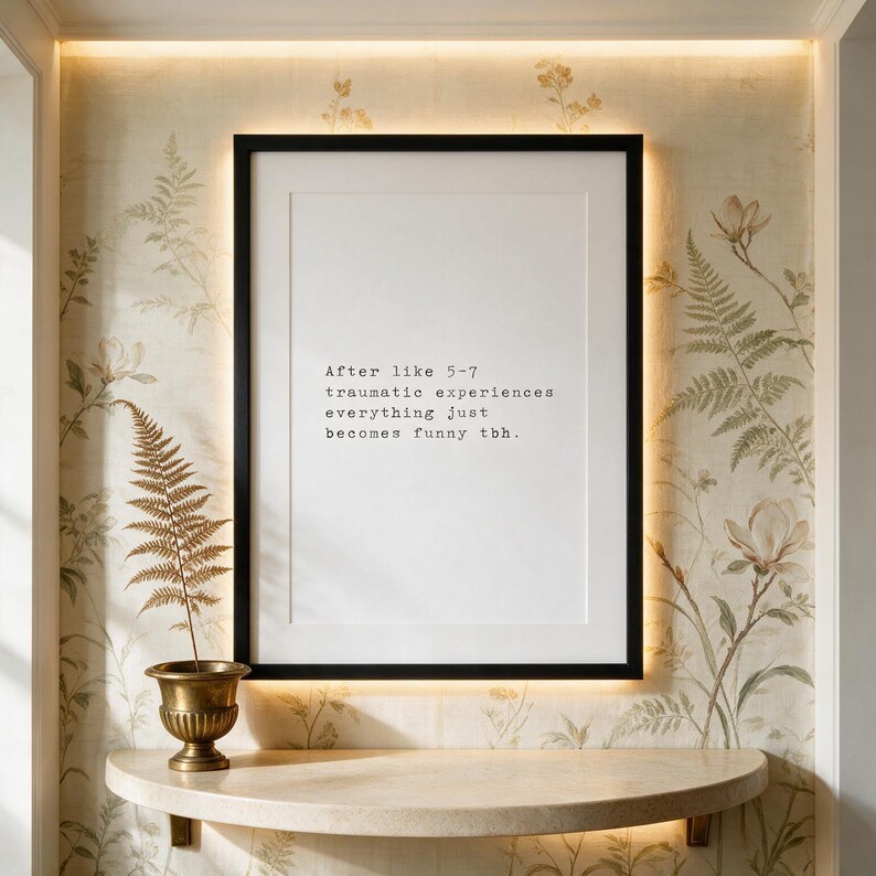 Framed black and white typographic dark humour print on a hand-painted botanical silk wall above a curved stone console.