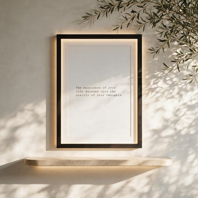 Framed black and white stoic quote print in a black wood frame with a warm LED halo, set above a pale stone shelf against textured plaster near olive branches, a considered gift for a calm home office.