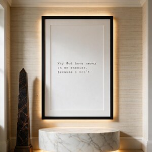 Framed black and white typographic dark humour print on a woven oyster silk wall above a round marble plinth.