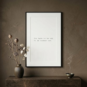 Op de afbeelding: Een ingelijste print met de tekst "You have to be odd to be number one." De zwart omlijste print wordt weergegeven op een donkerbruin oppervlak. Een vaas met gedroogde bloemen en een kleine kom staan ook op het oppervlak.