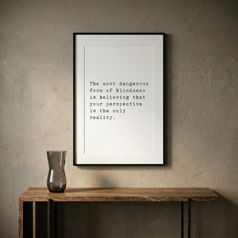 May include: A framed wall art print featuring the black text: The most dangerous form of blindness is believing that your perspective is the only reality. The print is displayed in a thin black frame with a white mat border on a textured wall.