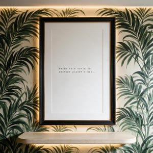 May include: A rectangular black frame containing a white print with the black typewriter-style text: "Maybe this world is another planet's hell." The frame is mounted on a wall with green palm leaf wallpaper and features a light-colored shelf below.