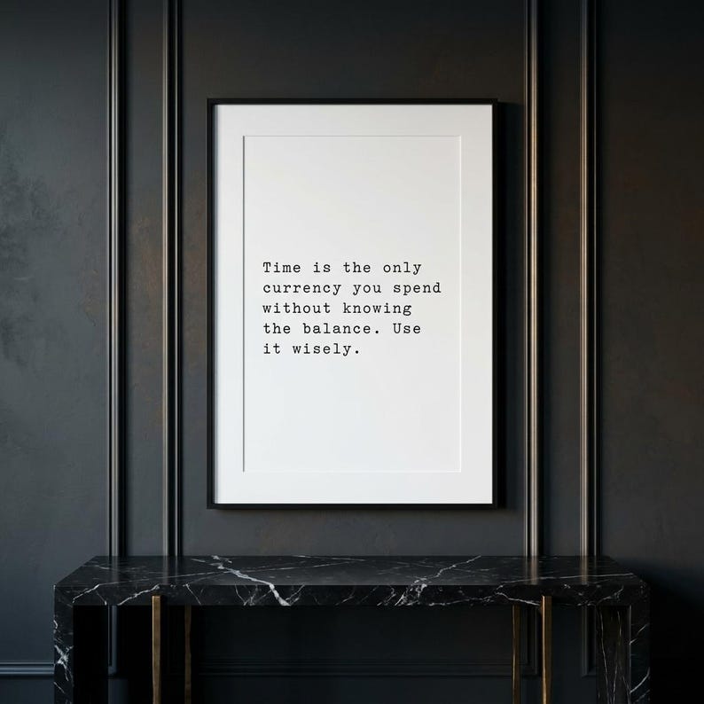 May include: A rectangular framed wall art piece featuring a white background with black typewriter-style text that reads: Time is the only currency you spend without knowing the balance. Use it wisely. The frame is thin and black.