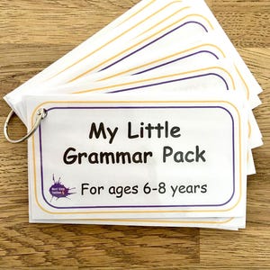 Może przedstawiać: Stos białych prostokątnych fiszek spiętych srebrnym metalowym kółkiem. Na wierzchniej karcie znajduje się czarny napis My Little Grammar Pack For ages 6-8 years, otoczony fioletowo-żółtą ramką oraz małe fioletowe logo w rogu karty.