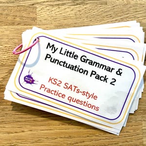 Peut inclure: Un paquet de fiches rectangulaires blanches reliées par un anneau métallique rose. La carte supérieure porte le texte My Little Grammar & Punctuation Pack 2, KS2 SATs-style Practice questions et un logo en forme d'éclaboussure d'encre violette.