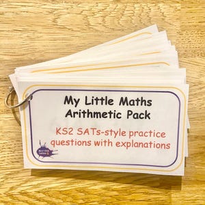 Può includere: Un mazzo di schede rettangolari bianche tenute insieme da un anello metallico. La scheda superiore riporta il testo My Little Maths Arithmetic Pack e KS2 SATs-style practice questions with explanations, con un piccolo logo nell'angolo.