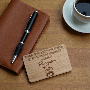 Könnte beinhalten: Eine Holzplakette mit dem Text "WENN PINGUINE IHREN PARTNER FINDEN, BLEIBEN SIE FÜR IMMER ZUSAMMEN. DU BIST MEIN Pinguin" und einer Pinguin-Illustration. Ein schwarzer Stift und ein braunes Leder-Notizbuch sind ebenfalls zu sehen.