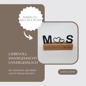 May include: A wooden sign with the letters "M" and "S" in black, with two intertwined heart shapes. The sign is on a wooden base, with the text "Hand in Hand - ein Leben lang" engraved. The dimensions are approximately 5.9 x 1.3 x 3 inches.