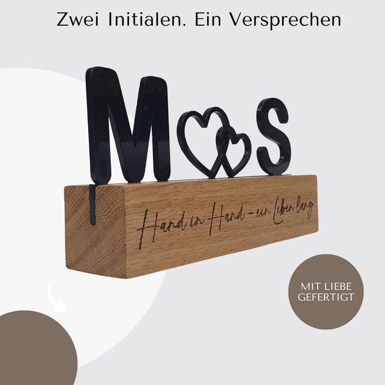 May include: A wooden block with the letters "M", a heart, and "S" in black. The block has the engraved text "Hand in Hand - ein leben lang". The words "Zwei Initialen. Ein Versprechen" are at the top. The words "MIT LIEBE GEFERTIGT" are at the bottom.