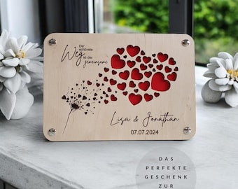 Könnte beinhalten: Holzschilder mit einem roten Herzdesign und dem Text "Der schönste Weg ist der gemeinsame" und "Lisa & Jonathan 07.07.2024". Das Schild hat auch ein Löwenzahndesign mit wegfliegenden Samen.