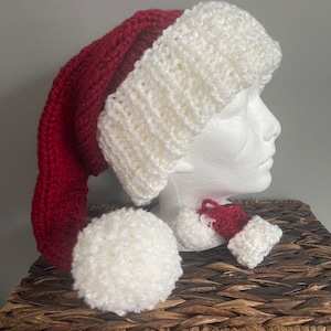May include: A red and white knitted Santa hat with a white pom-pom. The hat is on a mannequin head and there is a smaller white pom-pom on the table.