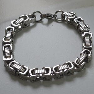 Puede incluir: Una pulsera plateada con un dise&ntilde;o de cadena de caja. La pulsera presenta eslabones rectangulares conectados por eslabones m&aacute;s peque&ntilde;os y redondeados. El cierre es un cierre simple de gancho y anillo. La pulsera est&aacute; hecha de un material met&aacute;lico brillante.