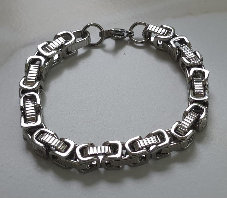 Puede incluir: Una pulsera plateada con un dise&ntilde;o de cadena de caja. La pulsera presenta eslabones rectangulares conectados por eslabones m&aacute;s peque&ntilde;os y redondeados. El cierre es un cierre simple de gancho y anillo. La pulsera est&aacute; hecha de un material met&aacute;lico brillante.