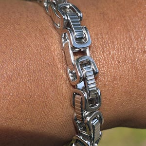 Puede incluir: Un brazalete plateado con un dise&ntilde;o de eslabones rectangulares. El brazalete est&aacute; hecho de metal brillante y se lleva en la mu&ntilde;eca. Los eslabones est&aacute;n conectados en un patr&oacute;n repetitivo, creando un aspecto audaz y moderno.