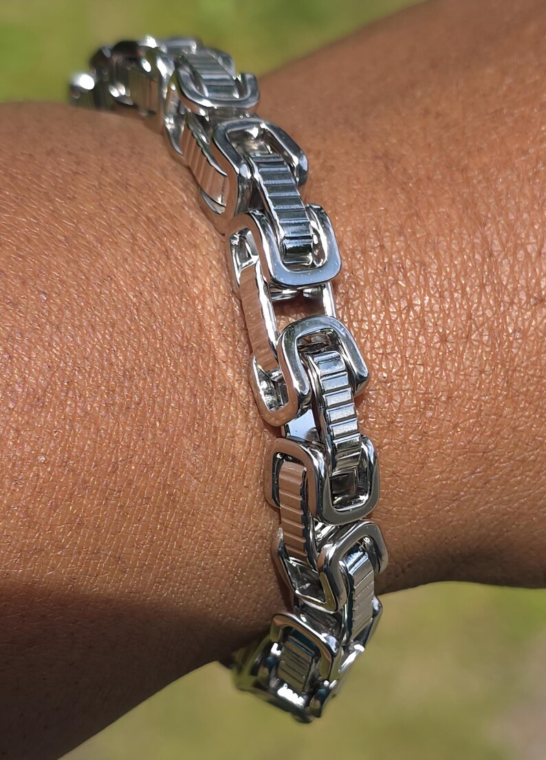Puede incluir: Un brazalete plateado con un dise&ntilde;o de eslabones rectangulares. El brazalete est&aacute; hecho de metal brillante y se lleva en la mu&ntilde;eca. Los eslabones est&aacute;n conectados en un patr&oacute;n repetitivo, creando un aspecto audaz y moderno.