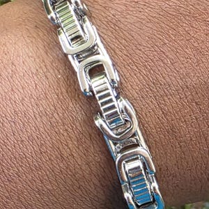 Puede incluir: Un brazalete plateado con una serie de eslabones rectangulares. Los eslabones est&aacute;n conectados por una serie de piezas curvas e interconectadas. El brazalete se lleva en la mu&ntilde;eca.