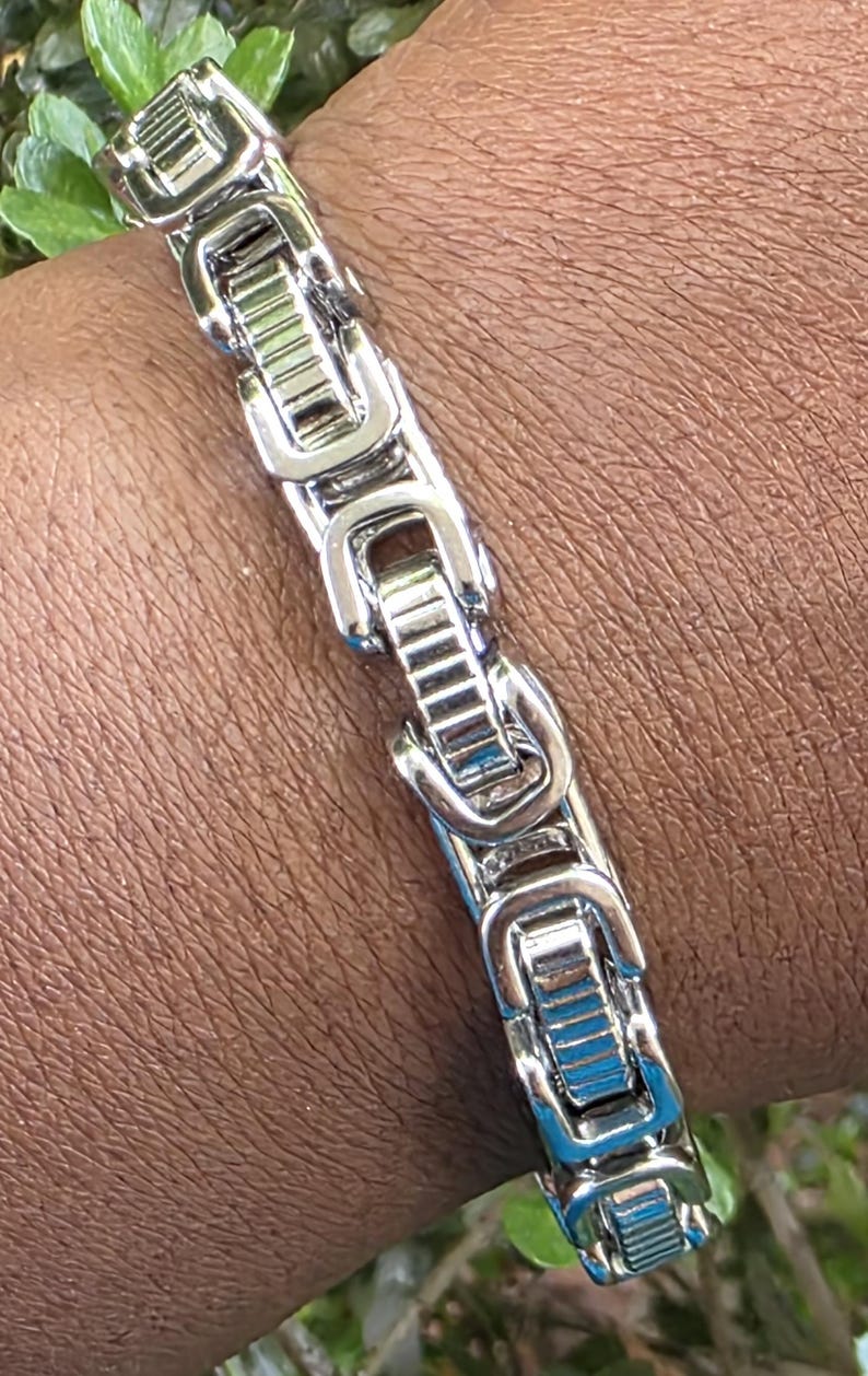 Puede incluir: Un brazalete plateado con una serie de eslabones rectangulares. Los eslabones est&aacute;n conectados por una serie de piezas curvas e interconectadas. El brazalete se lleva en la mu&ntilde;eca.