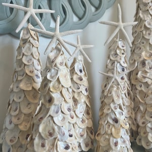 May include: Several decorative seashell Christmas trees topped with white starfish. The trees are made of small, cream-colored shells and are arranged on a light blue surface. A light blue, decorative mirror is in the background.