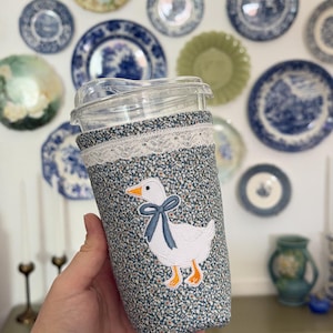May include: A reusable coffee cup sleeve with a floral pattern in shades of blue and green. The sleeve has a white lace trim and an embroidered goose with a blue bow. The cup is clear with a plastic lid.