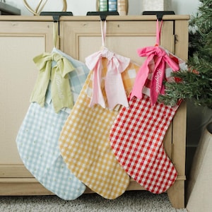 May include: Three Christmas stockings in pastel shades hang from a wooden cabinet. The stockings display gingham patterns in blue, yellow, and red. Each stocking has a matching ribbon bow with an embroidered name.