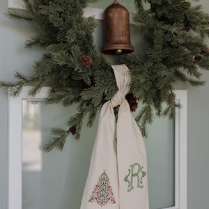 Peut inclure: Un lien en tissu couleur crème avec un sapin de Noël brodé en vert et rouge à une extrémité et une lettre R brodée en vert à l'autre. Le lien est noué et suspendu à une couronne de pin verte avec de petites pommes de pin brunes et une cloche en bois.