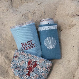 Op de afbeelding: Drie stoffen drankhouders en een kleine etui liggen op zand. Eén houder is lichtblauw met verticale strepen en de geborduurde tekst "EAST COAST". Een andere is zeegroen met een wit schelpmotief. De etui heeft een bloemenpatroon en een rode kreeft.