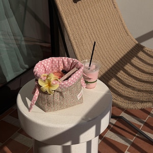 May include: A small, round side table holds a pink-and-white polka-dotted basket with a yellow flower and a pink drink in a clear plastic cup. The basket is filled with items. A woven chair is in the background.