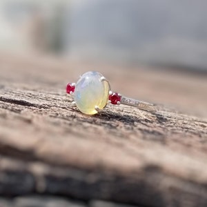 May include: A silver ring with a white opal gemstone and two small red ruby accents.