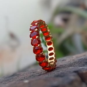 May include: A gold ring featuring a continuous line of oval, deep red gemstones. The ring is set against a blurred natural backdrop, accentuating the vibrant colour of the stones. The ring is a piece of jewellery.