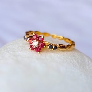 May include: A gold ring with a flower design. The flower is made of red and black gemstones, with a single white gemstone at its centre. The ring's band has a textured, beaded pattern.
