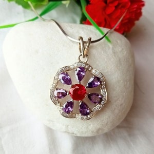May include: A silver pendant necklace with a red and purple flower design. The pendant features a red gemstone in the center surrounded by purple gemstones.