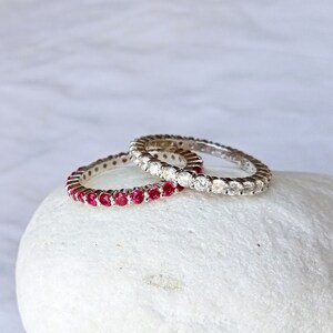 May include: Two eternity bands, one with red gemstones and one with clear gemstones, on a white rock.