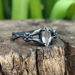 May include: A black metal ring with a clear, marquise-cut gemstone. The ring has floral and leaf detailing on the band. The ring is set on a rustic wooden surface, with a blurred green background.